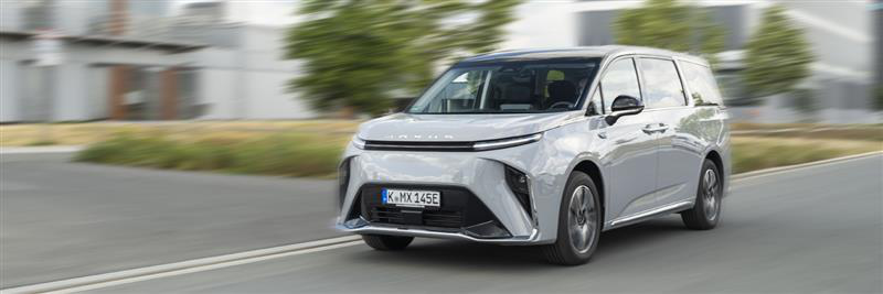 Ein silberner Elektro-Minivan fährt auf einer Landstraße mit Bäumen im Hintergrund.